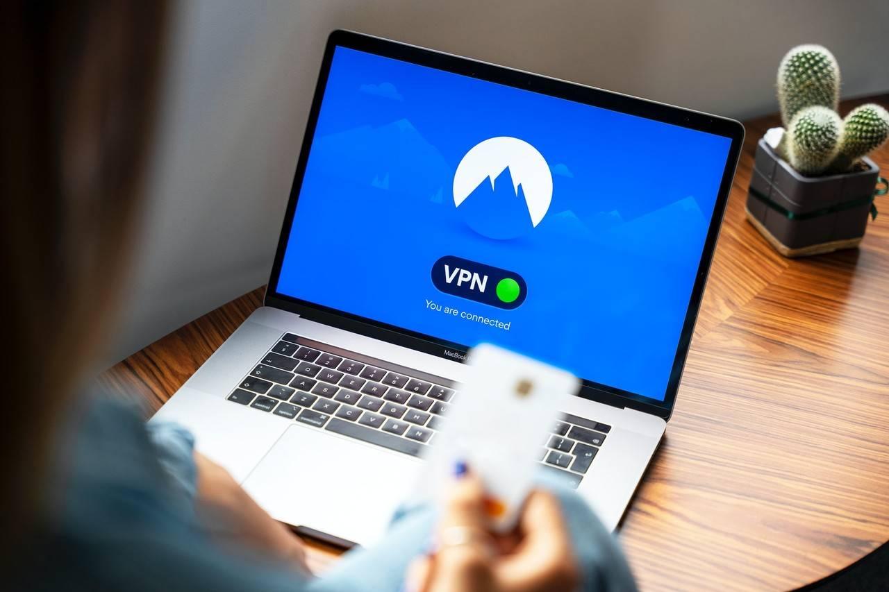 Laptop mostrando uma tela azul com a mensagem "VPN - Você está conectado".  Uma pessoa sentada atrás do laptop, parcialmente visível.