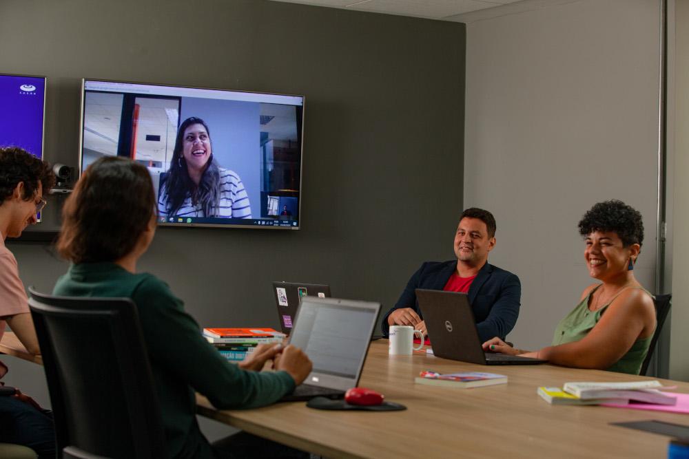 Várias pessoas reunidas em uma mesa numa sala de reunião, com uma pessoa participando pela TV
