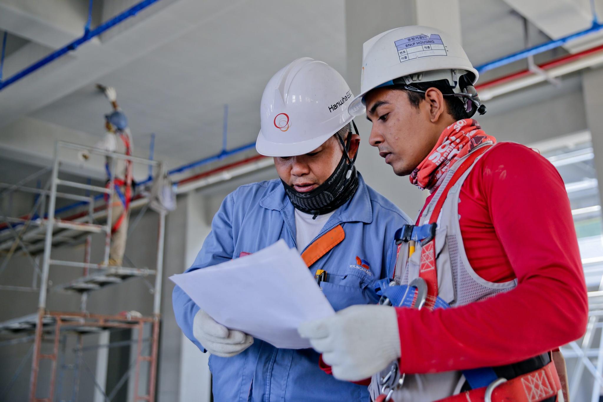 Dois trabalhadores da construção civil examinam um documento em conjunto, usando capacetes de segurança e equipamentos de proteção individual. Um trabalhador de andaime está ao fundo, fora de foco.