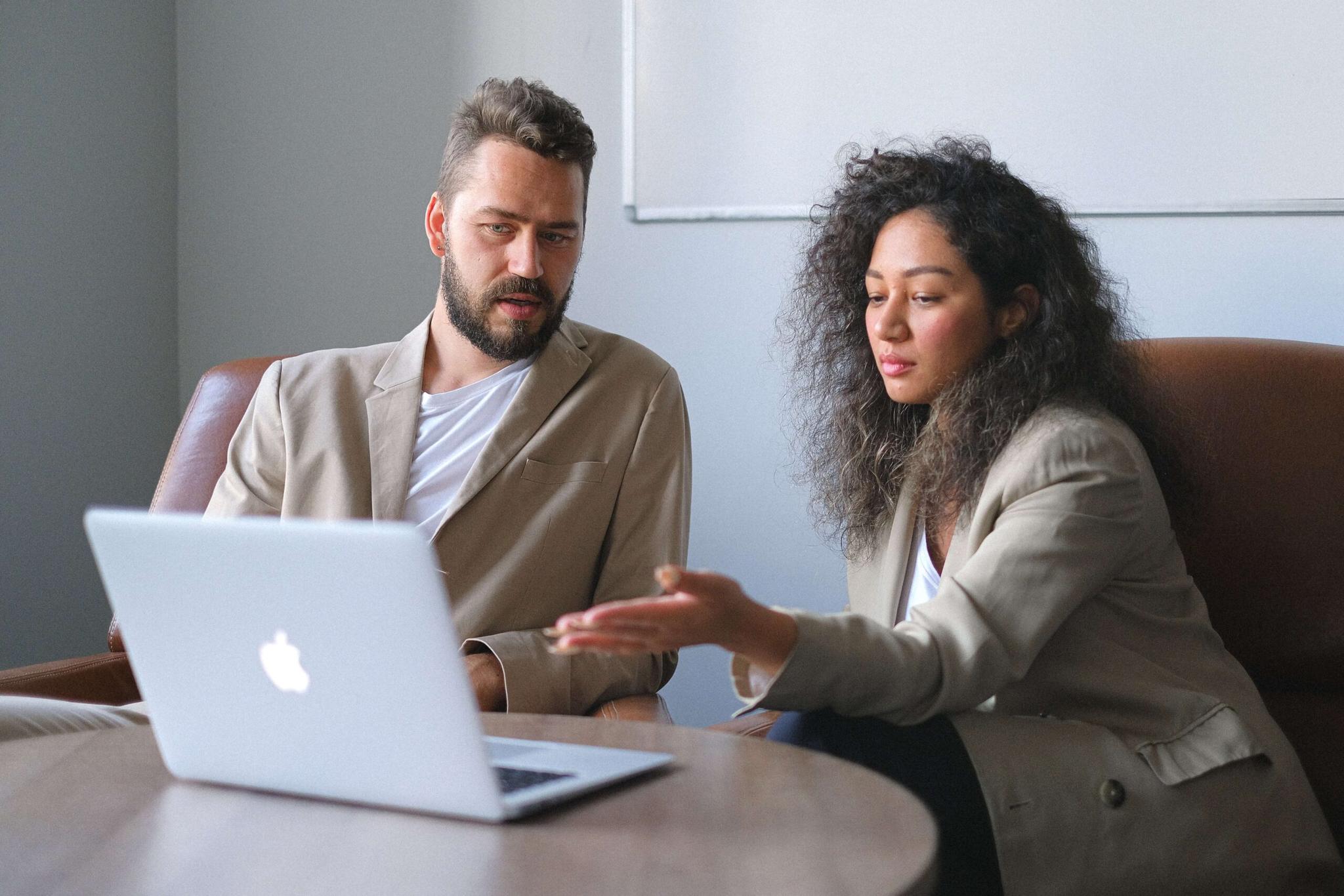 Um homem e uma mulher, ambos vestindo blazers bege, sentados em poltronas de couro e analisando um laptop.  A mulher está gesticulando enquanto eles discutem algo na tela.