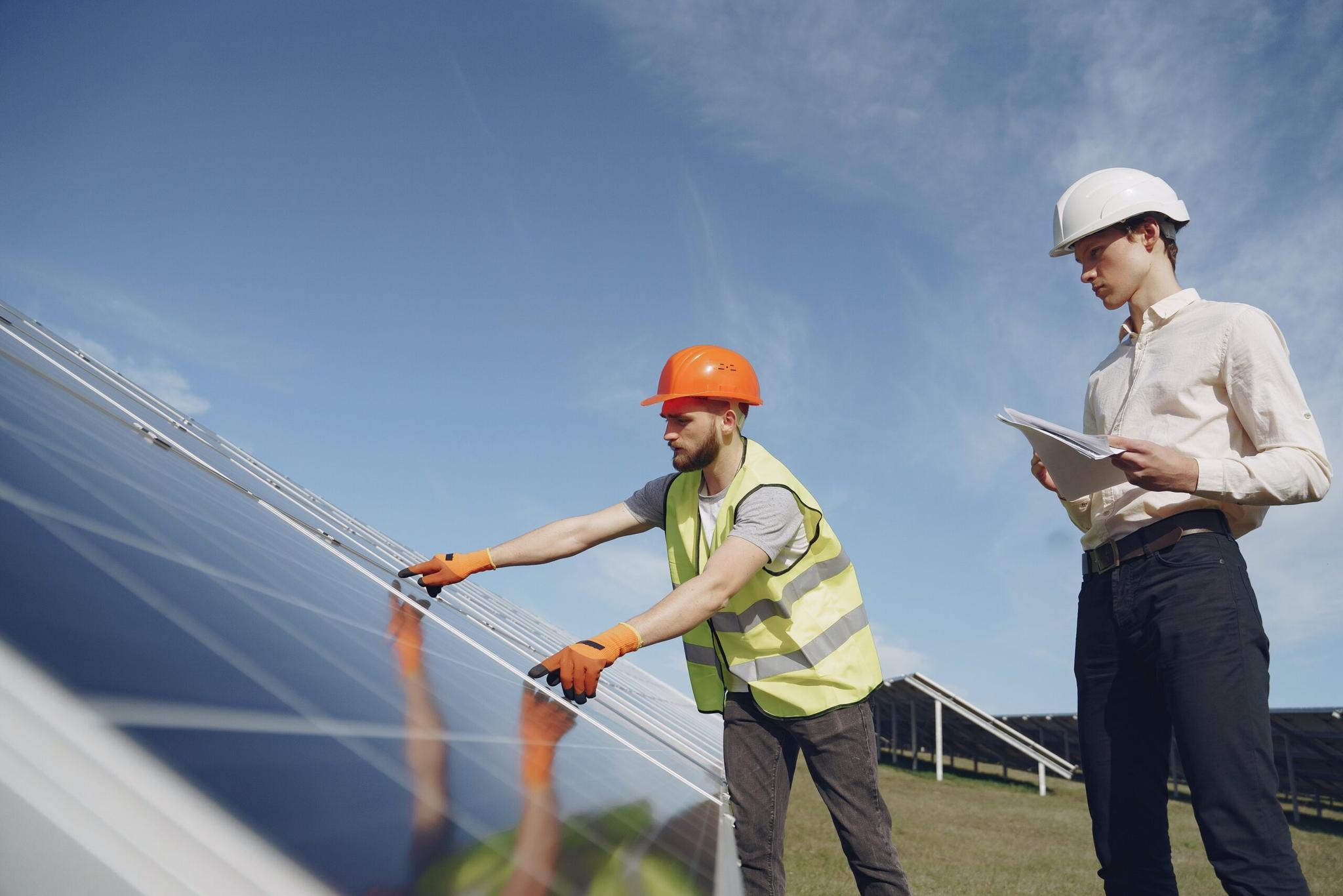 Dois trabalhadores inspecionam painéis solares em um campo ensolarado. Um deles usa um colete de segurança e luvas de trabalho, enquanto o outro revisa papéis.
