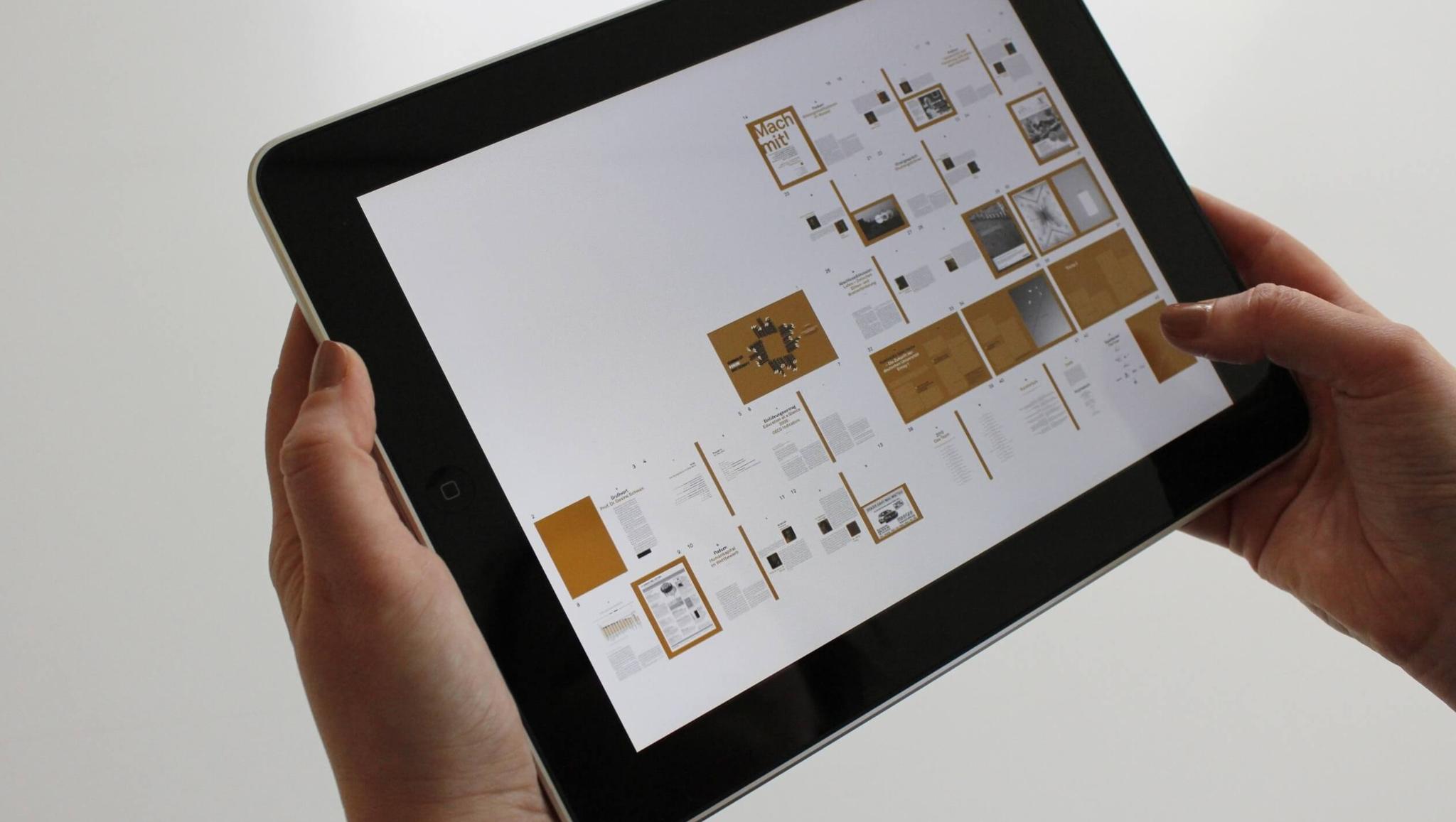 Duas mãos segurando um tablet que exibe uma publicação digital com páginas em miniatura em um layout de grade.