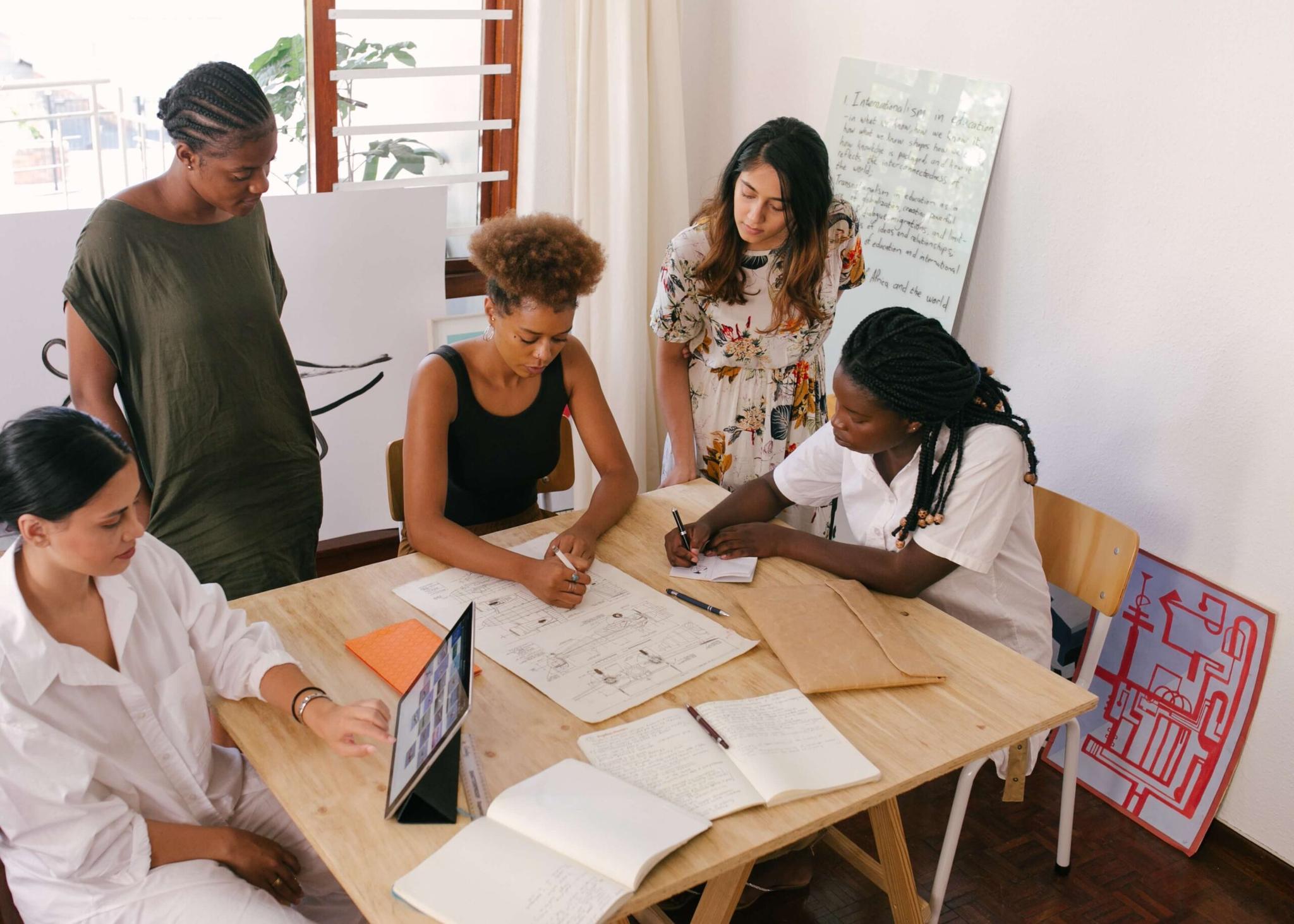 Quatro mulheres colaboram em uma mesa de madeira, analisando um projeto e tomando notas em cadernos.  Uma mulher está usando um tablet.