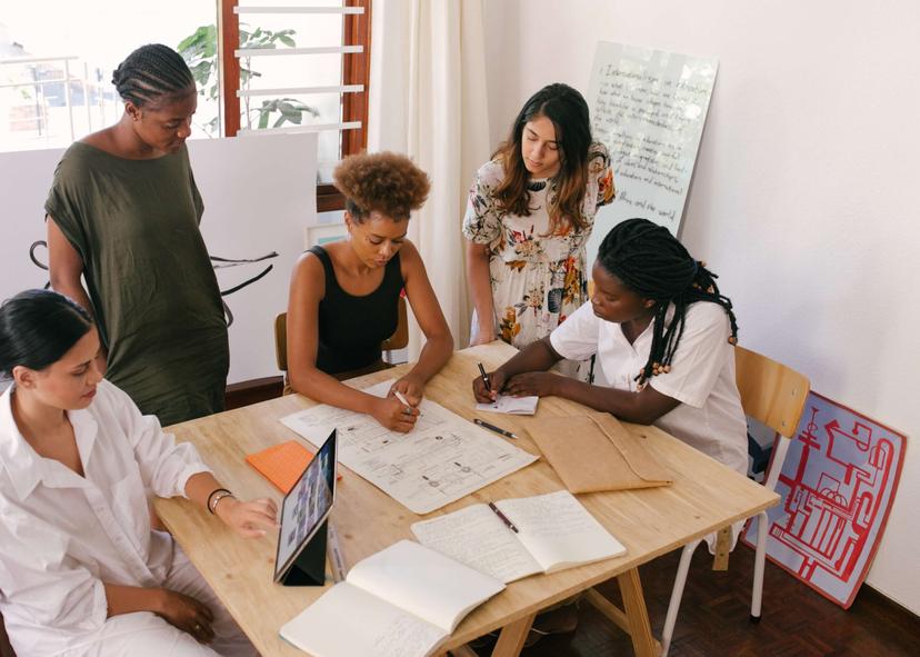 Quatro mulheres colaboram em uma mesa de madeira, analisando um projeto e tomando notas em cadernos.  Uma mulher está usando um tablet.