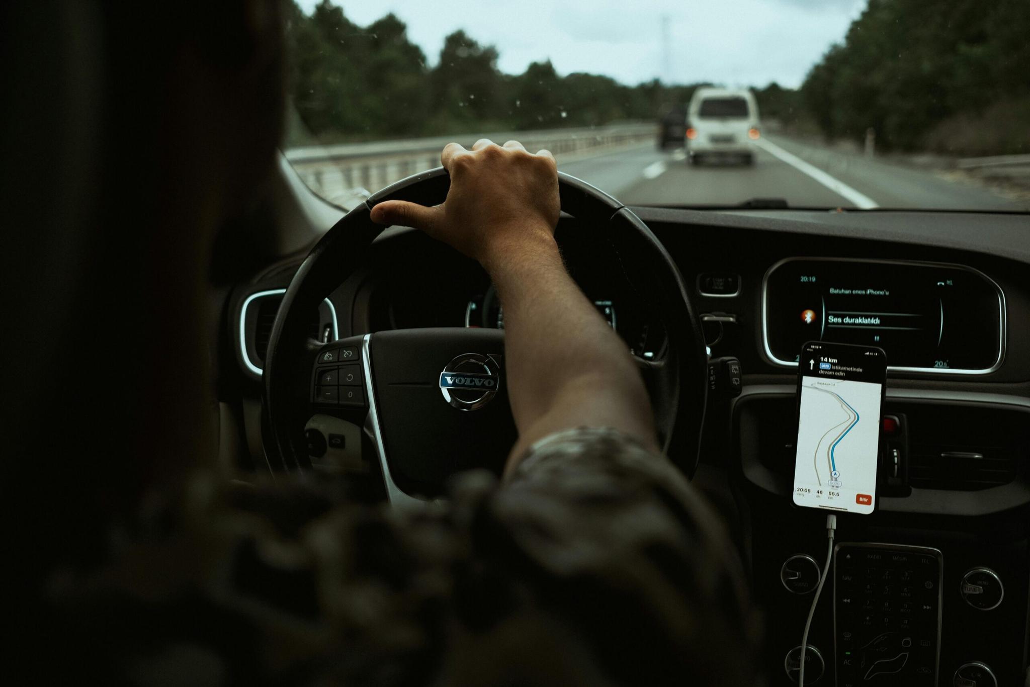 Visão em primeira pessoa de um condutor numa Volvo, segurando o volante, com um smartphone com GPS no painel. A estrada está visível através do para-brisa.