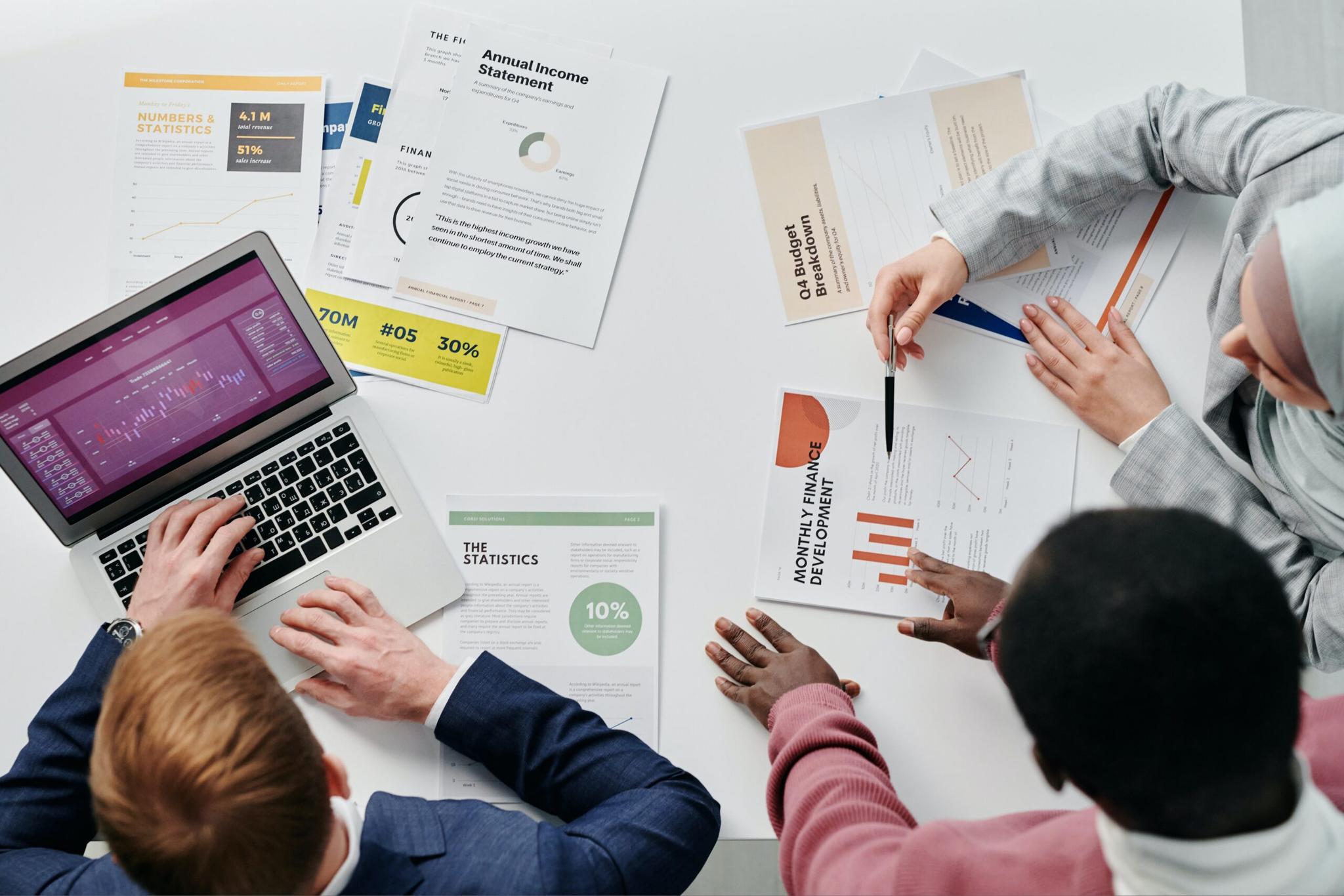 Vista aérea de três colegas analisando relatórios financeiros e gráficos em uma mesa de escritório.
