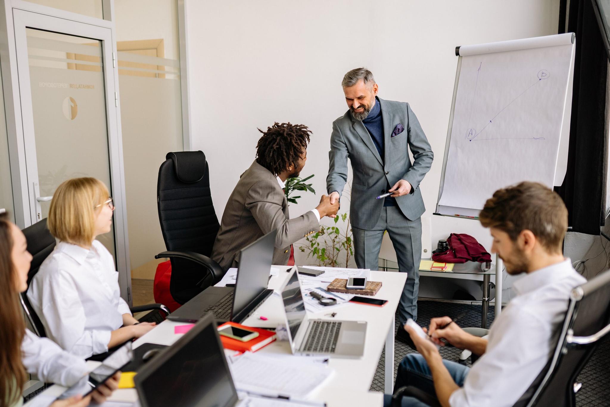 Reunião de negócios em um escritório moderno, com dois homens apertando as mãos enquanto outros colegas assistem a uma apresentação em um quadro branco.