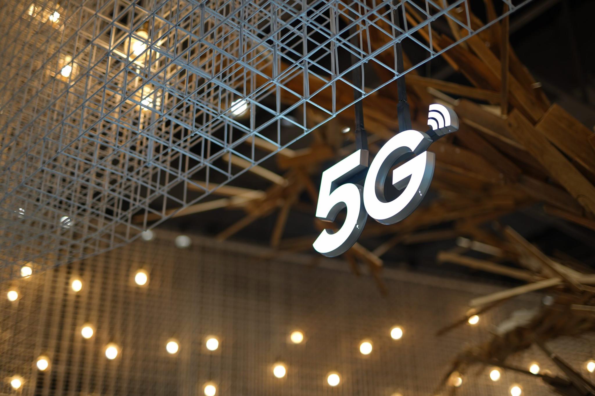 Um ícone em LED da logo "5G", pendurado em uma estrutura e ao fundo uma árvore e algumas lâmpadas.