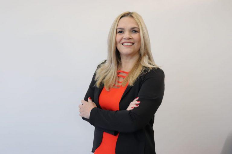 Uma mulher loira sorridente, usando um blazer preto sobre um vestido vermelho, com os braços cruzados.