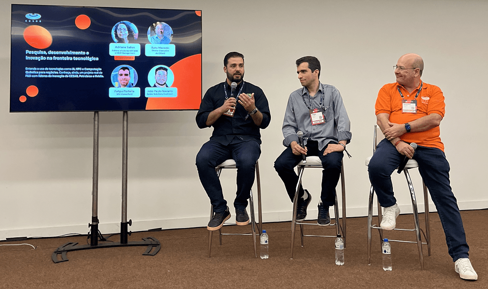 Três palestrantes sentados em banquinhos, participando de um painel em um evento tecnológico. Um telão atrás deles exibe informações sobre pesquisa, desenvolvimento e inovação.