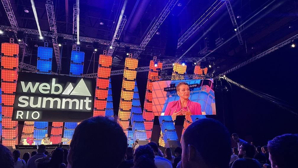 Uma palestrante no palco do Web Summit Rio, exibida em uma tela grande, com uma grande plateia. Iluminação vibrante e estrutura de palco colorida em cubos.