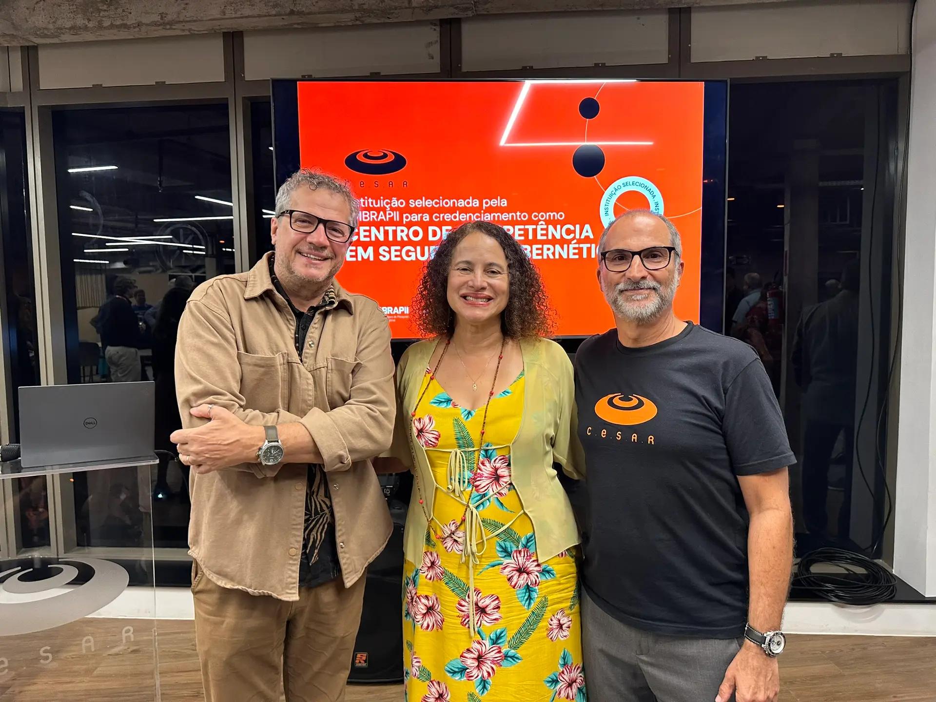 Três pessoas posam sorrindo para uma foto em frente a uma tela com um anúncio de credenciamento.