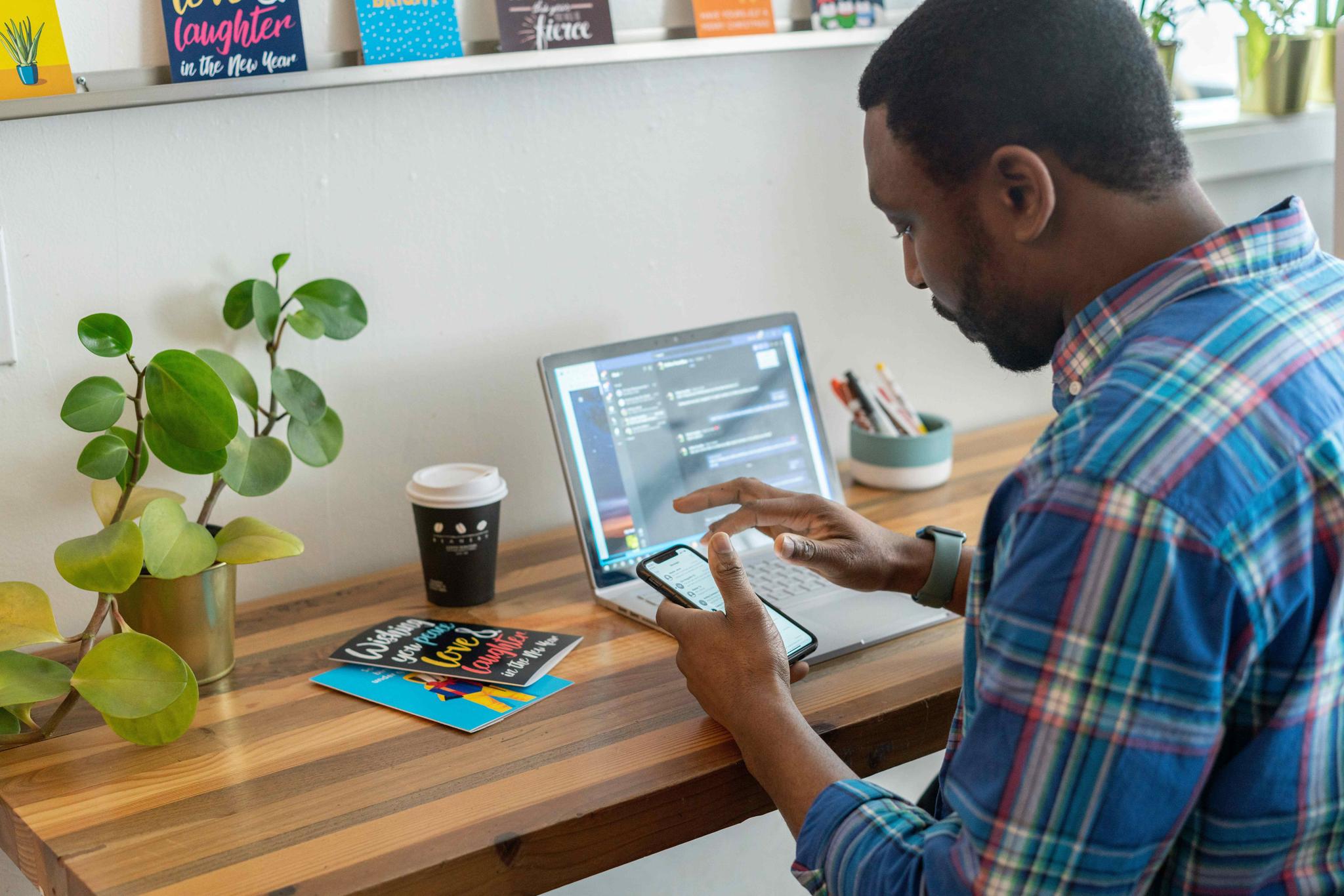Home office scene: A man in a plaid shirt sits at a wooden desk, using his smartphone and laptop. Plants and greeting cards are visible on the desk.