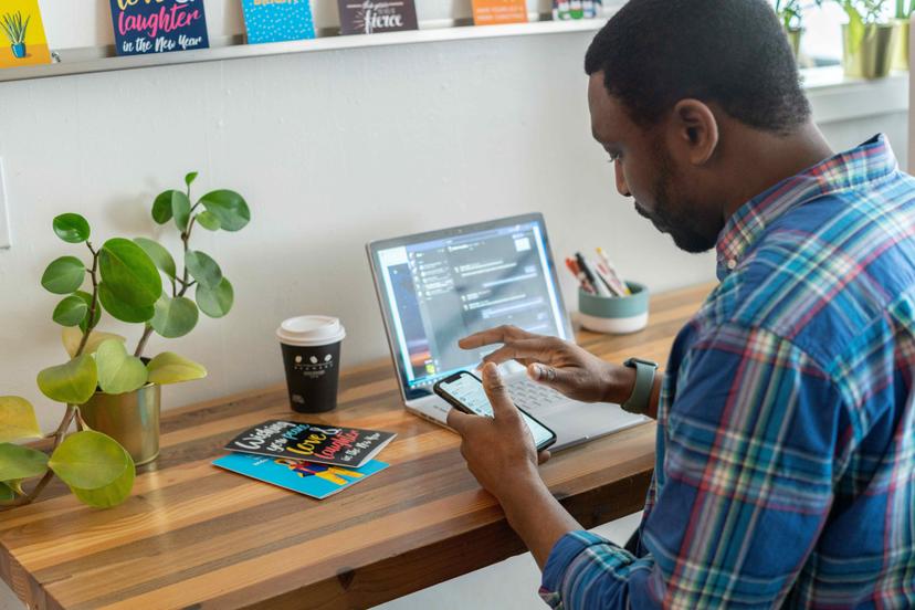 Home office scene: A man in a plaid shirt sits at a wooden desk, using his smartphone and laptop. Plants and greeting cards are visible on the desk.
