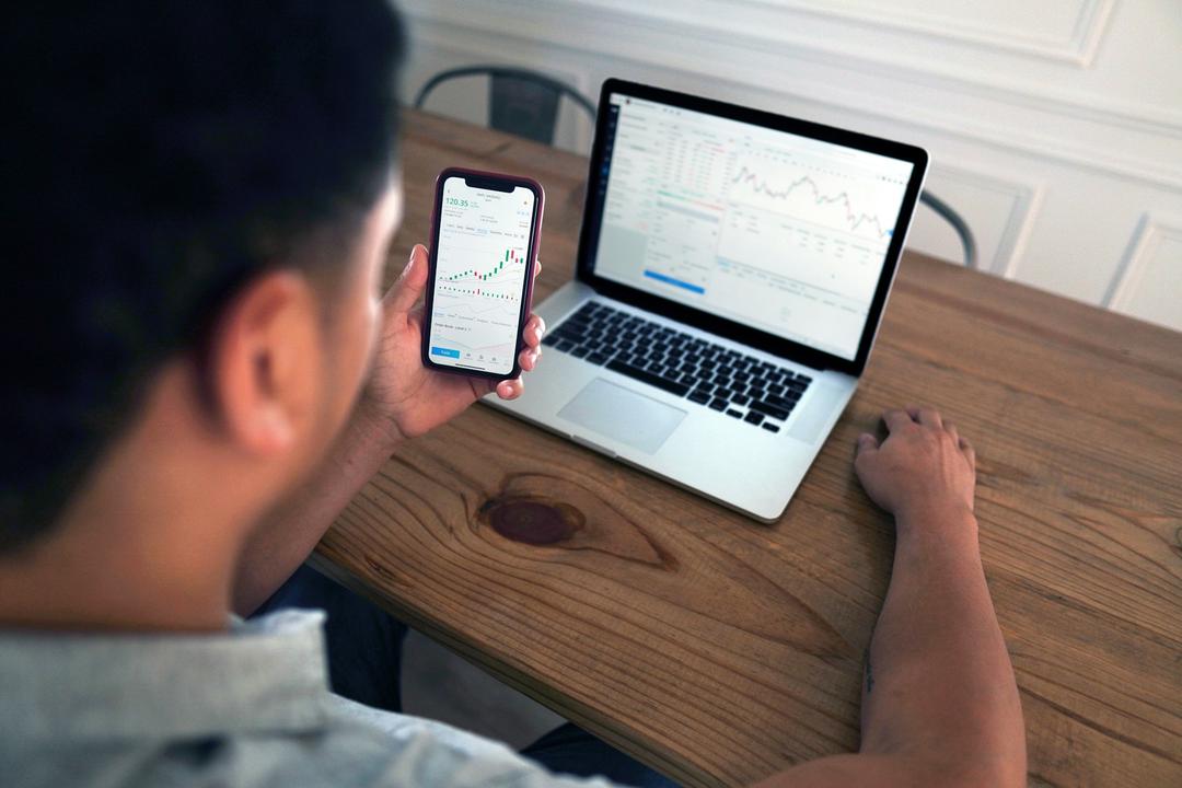 Pessoa analisa gráficos financeiros em smartphone e notebook sobre mesa de madeira