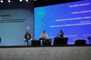 Painel de discussão em uma conferência com três palestrantes no palco, apresentando um fundo azul e ícones de segurança.