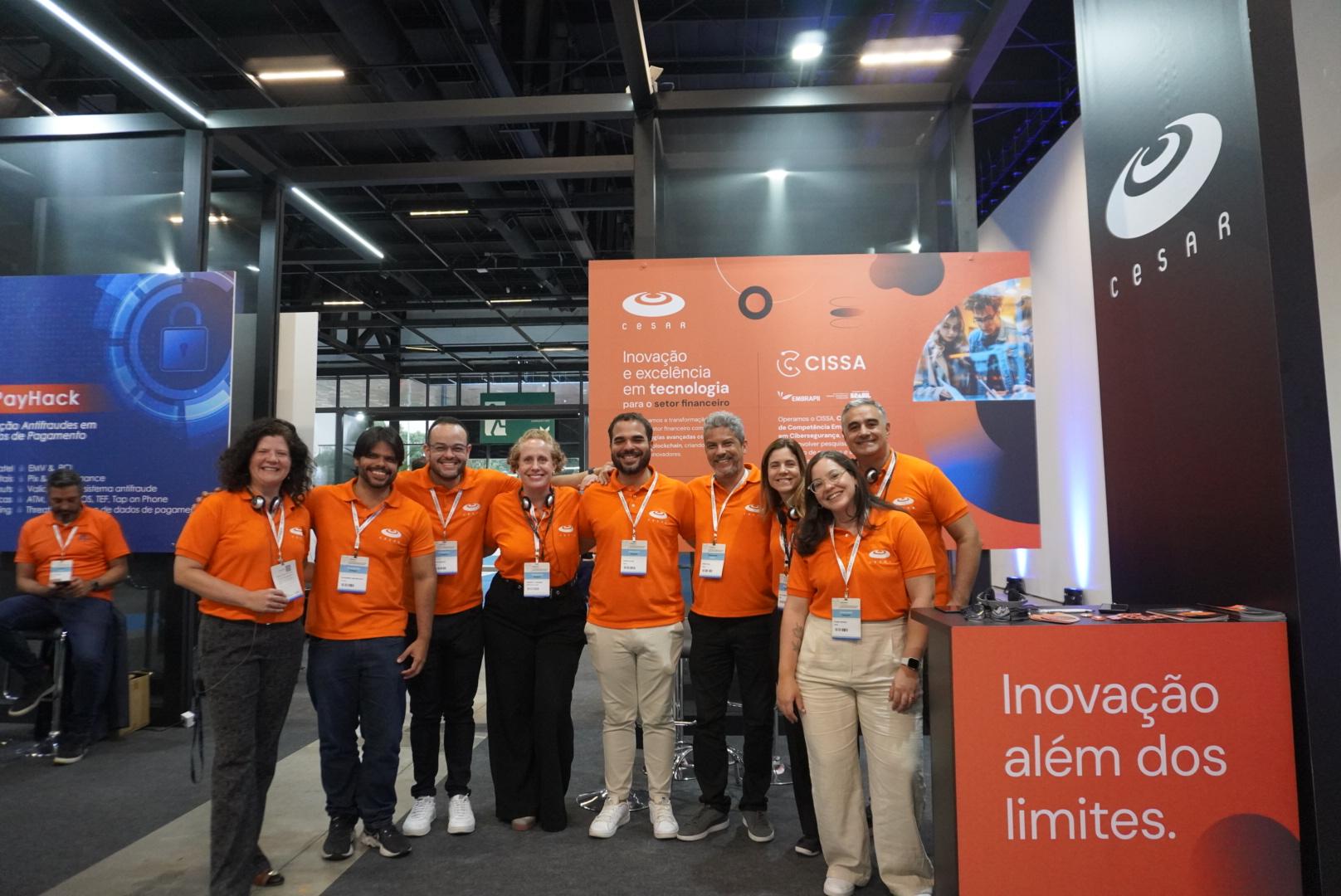 Equipe em uniforme laranja posa para foto em evento de tecnologia financeira.  Banner com logotipos CESAR e CISSA ao fundo.