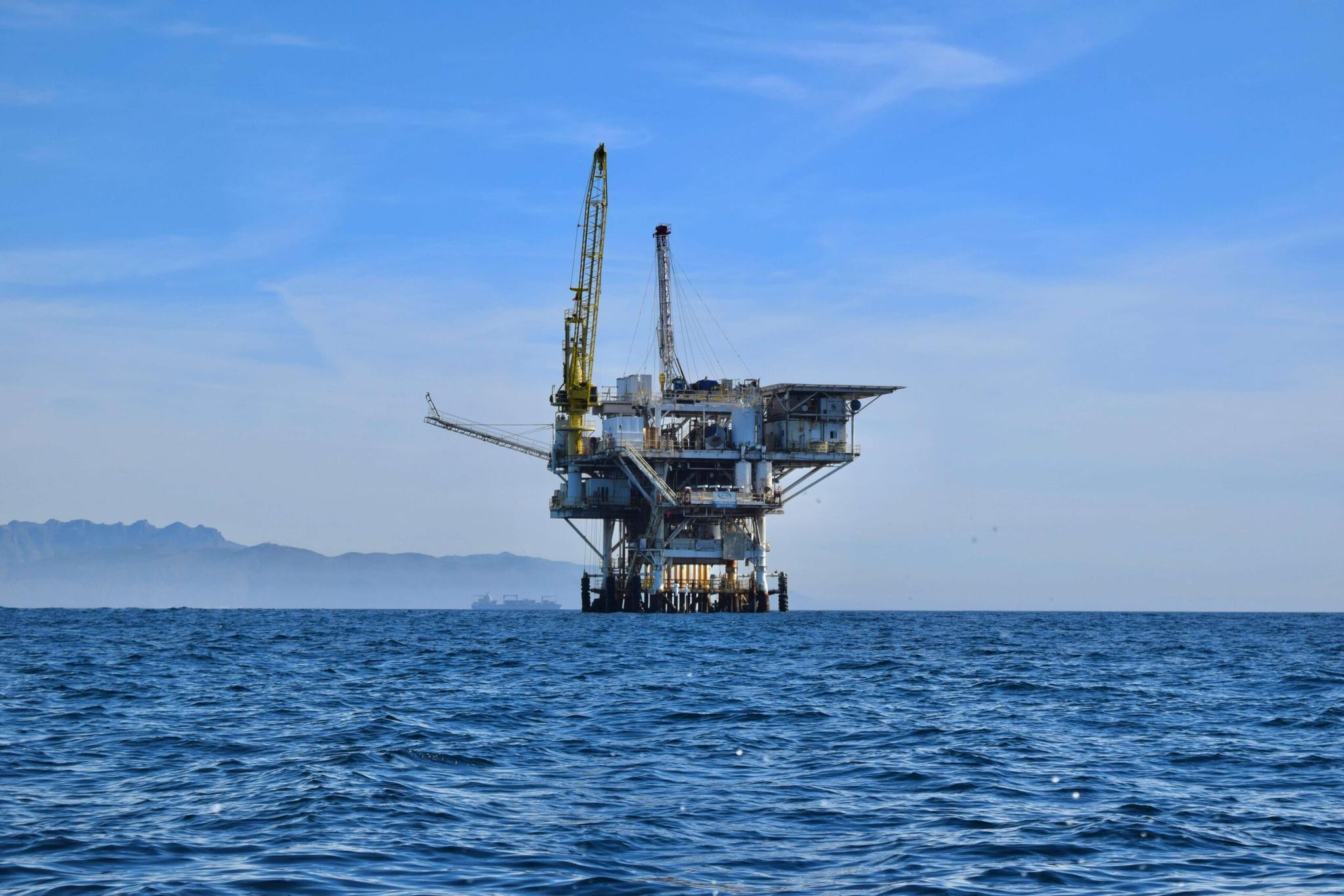 Plataforma petrolífera em alto-mar sob um céu azul claro, com montanhas ao fundo.