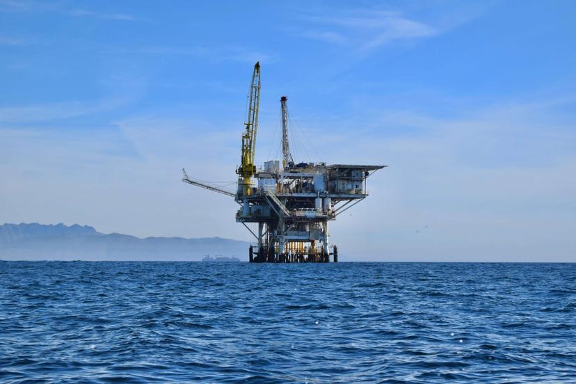 Plataforma petrolífera em alto-mar sob um céu azul claro, com montanhas ao fundo.