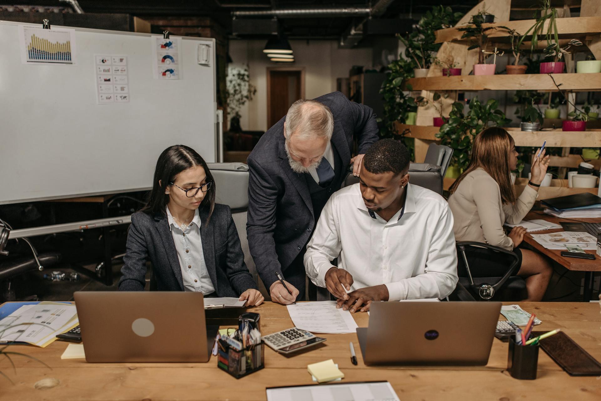 Três colegas de trabalho revisam documentos em uma mesa de escritório. Um homem mais velho está orientando um homem e uma mulher mais jovens. Um colega de trabalho adicional está sentado no fundo, trabalhando em seu computador.