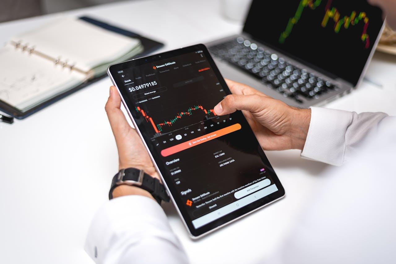 Close-up of a person's hands holding a tablet displaying a cryptocurrency stock chart.  A laptop with a similar chart is visible in the background.