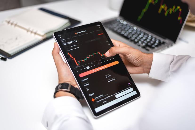 Close-up of a person's hands holding a tablet displaying a cryptocurrency stock chart.  A laptop with a similar chart is visible in the background.