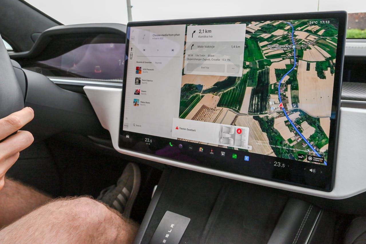 Close-up of a Tesla's central touchscreen displaying navigation, music options, and a seatbelt reminder; driver's legs and hand on the steering wheel are partially visible.