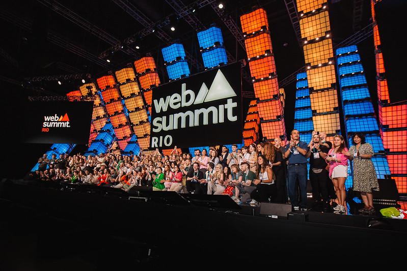 Grande grupo de pessoas aplaudindo em um palco com o letreiro "Web Summit Rio" e uma instalação de cubos coloridos ao fundo.