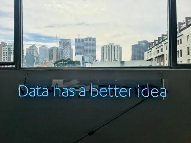 Data-driven e jornada analítica nas empresas Letreiro de neon azul com a frase "Data has a better idea" fixado abaixo de uma janela com vista para prédios