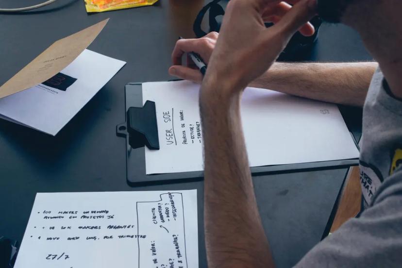 Experiência do usuário e como a pesquisa de design ajuda a criar soluções Homem apoia a cabeça com a mão enquanto escreve anotações com o título "USER SIDE" em uma prancheta sobre a mesa de trabalho