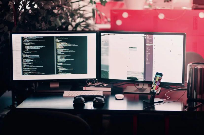 Futuro do trabalho e novas demandas do mercado tecnológico Dois monitores acesos mostram códigos e sistemas em uma mesa com teclado, mouse, fone de ouvido e celular