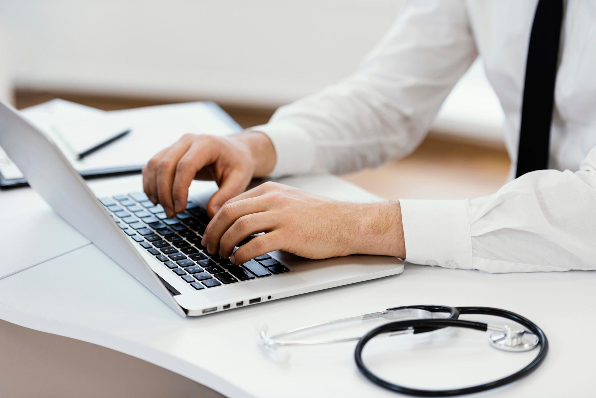 Ponto-care – Inovação em exames essenciais ao paciente em Nova Iorque Médico de camisa branca e gravata preta digita no computador portátil. Um estetoscópio preto repousa sobre a mesa branca.