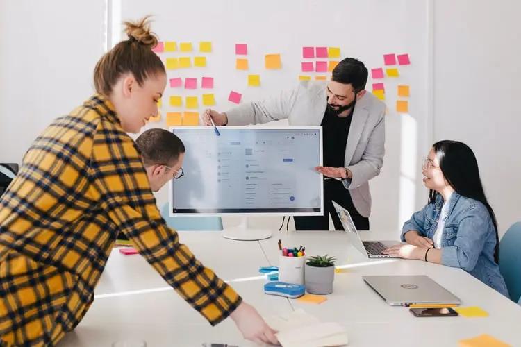 Design estratégico – entenda por que não é só sobre desenhos Quatro pessoas debatem ideias ao redor de uma mesa com notebooks e uma grande tela, diante de um painel de post-its