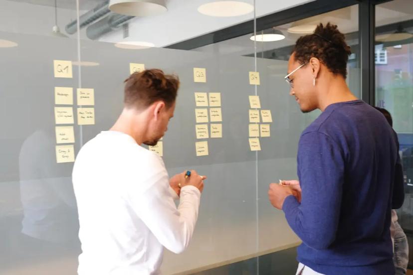 Jornada de inovação – descubra em qual etapa sua empresa está Dois homens trabalham organizando notas autoadesivas amarelas em uma parede de vidro divisória de escritório