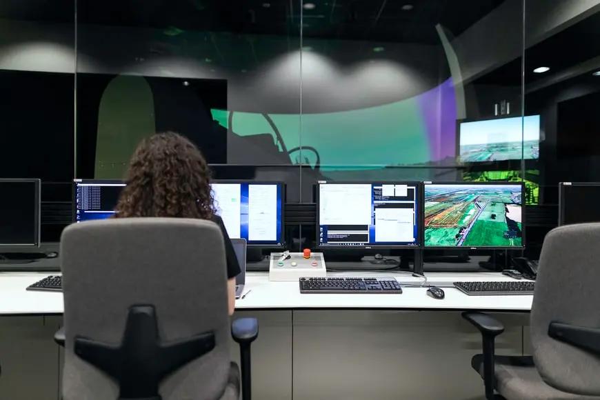 AIoT – o que é e como essa combinação tecnológica impacta negócios Profissional de costas monitora múltiplos computadores em uma sala de controle com uma grande tela de vidro ao fundo