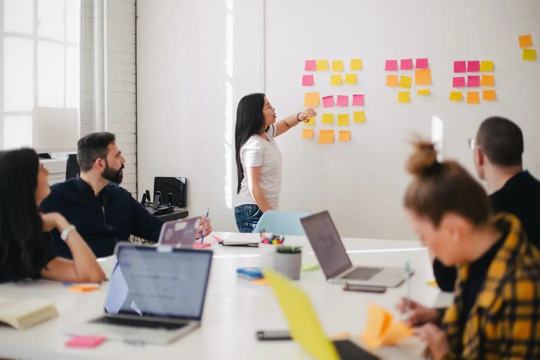 Liderança na inovação – saiba como construir uma cultura inovadora Mulher aponta para notas autoadesivas em uma parede branca durante reunião com equipe ao redor de mesa com notebooks