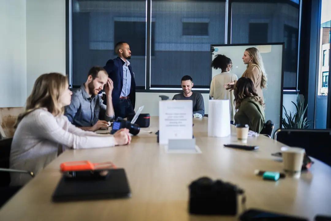Projetos de inovação e a importância de um olhar holístico no negócio Grupo de profissionais trabalha integrado em uma sala de reuniões ao redor de uma grande mesa de madeira com laptops
