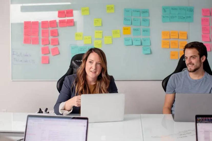 Product discovery – como o design ajuda a descobrir novos produtos de sucesso Uma mulher de blazer azul fala enquanto usa um notebook ao lado de um colega em uma mesa de escritório, com uma grande lousa branca cheia de post-its coloridos ao fundo