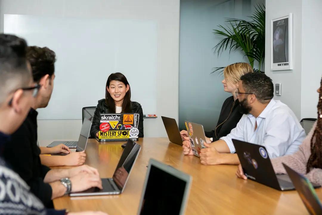 Liderança do futuro – 4 características essenciais para uma gestão melhor Uma mulher de casaco preto fala na cabeceira de uma mesa de reuniões cercada por profissionais com seus respectivos notebooks