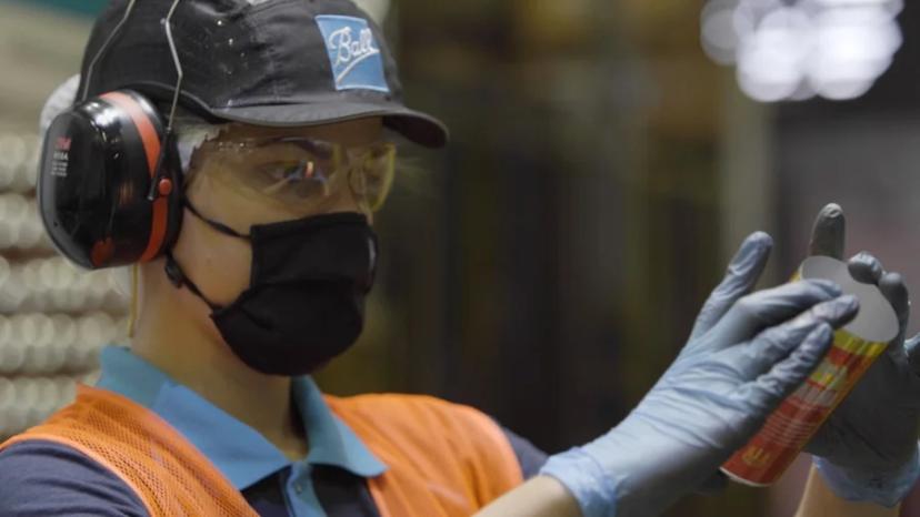 Manutenção preditiva e eficiência na indústria com a Ball e o CESAR Colaboradora de máscara, óculos de proteção e luvas azuis inspeciona detalhadamente uma lata de alumínio em linha de produção.