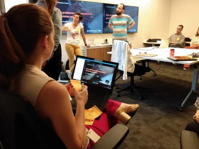 KC utiliza prototipação e validação em plataforma sobre menstruação Mulher em primeiro plano segura post-its diante de portátil. Ao fundo, equipe debate junto a ecrãs com slides de projeto