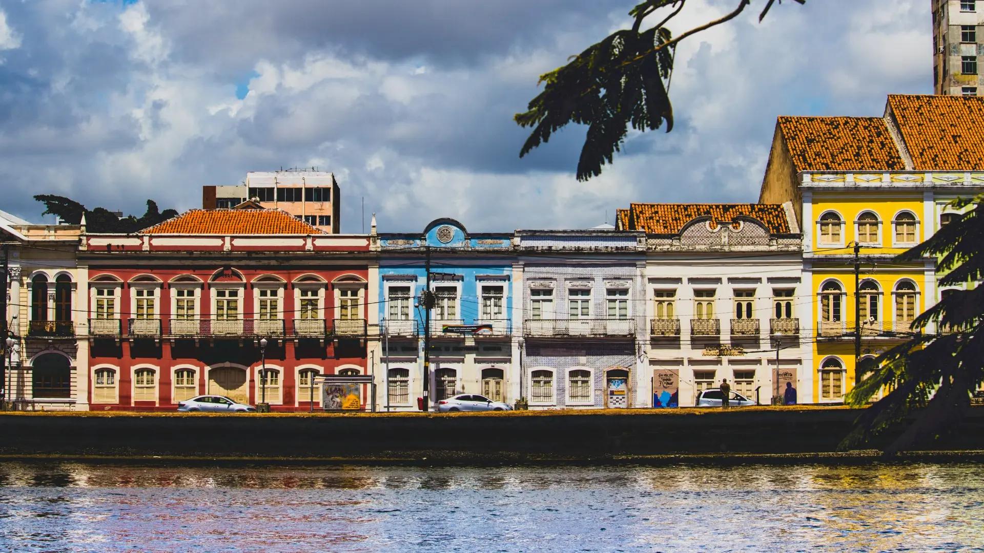 Design Thinking no turismo criativo em Recife e inovação cultural Casarões históricos coloridos na beira do rio no Recife Antigo sob céu nublado, vistos por trás de folhagens.