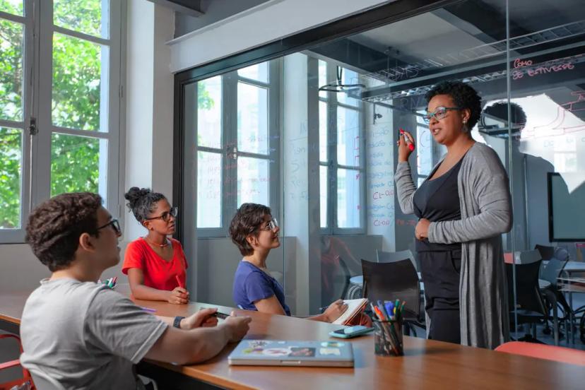 Educação corporativa e residência em TI para empresas Mulher explica conteúdo escrevendo em parede de vidro enquanto três jovens assistem sentados ao redor de uma mesa de madeira.