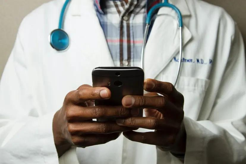 Programa de residência para desenvolvedores em tecnologia da saúde Médico negro em jaleco branco e estetoscópio azul usa smartphone para acessar dados e plataformas digitais de saúde.