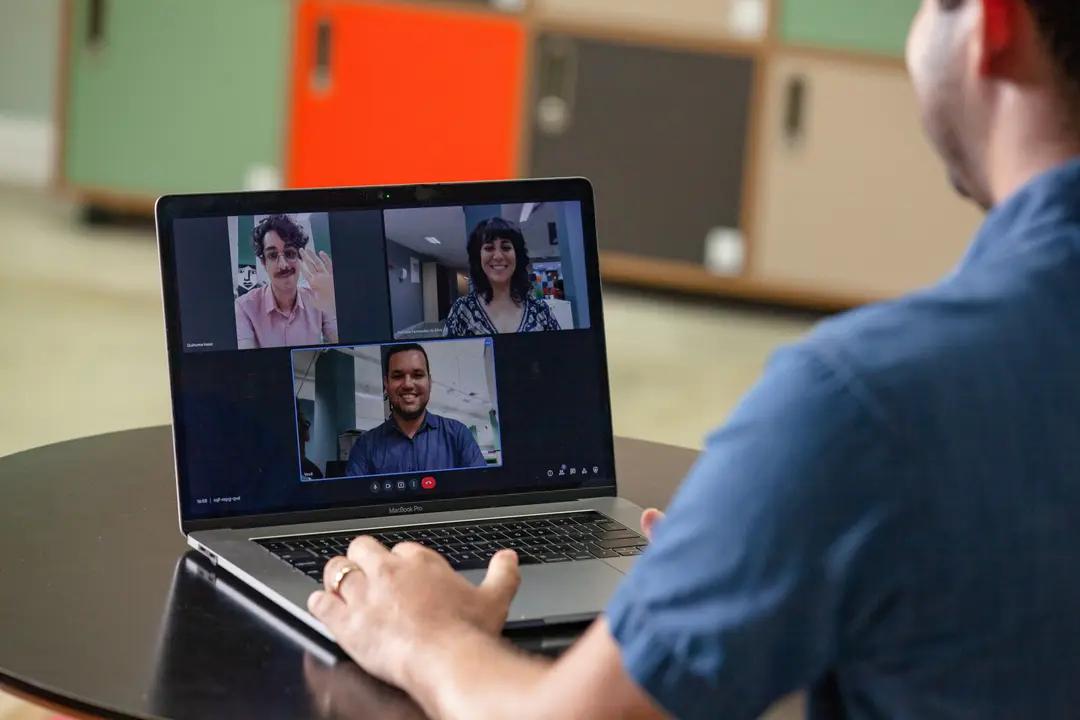 Liderança em design no contexto remoto e gestão de equipes ágeis Homem de costas faz videoconferência em laptop com três pessoas sorridentes na tela, sentados em frente a armários coloridos.