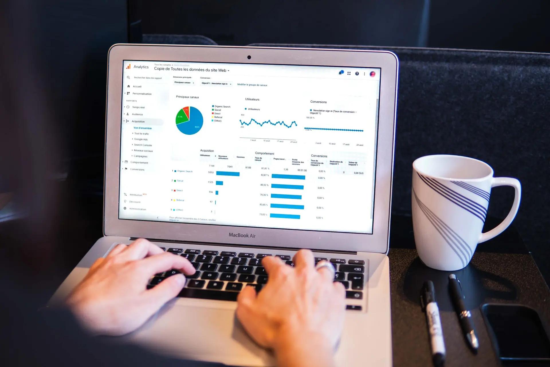 Data Analytics – a inteligência por trás da interpretação de dados digitais Pessoa vista de costas digita em um laptop que exibe gráficos de pizza e barras em um painel do Google Analytics, ao lado de uma caneca branca.