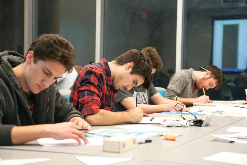 Aprendizagem no fluxo de trabalho Quatro homens jovens sentados lado a lado em uma longa mesa de escritório, concentrados enquanto escrevem e desenham em papéis instalados à sua frente