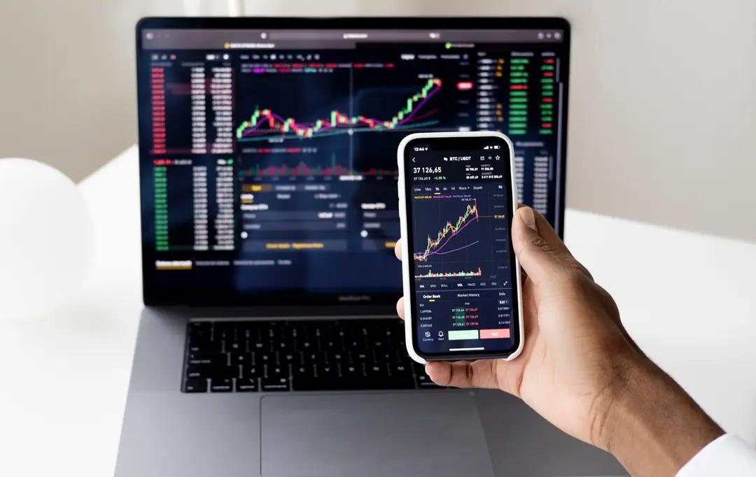 DeFi – finanças descentralizadas e seu impacto no sistema tradicional Mão segura um smartphone que exibe um gráfico de velas financeiras do par BTC/USDT em frente à tela de um laptop com gráficos de trade abertos.