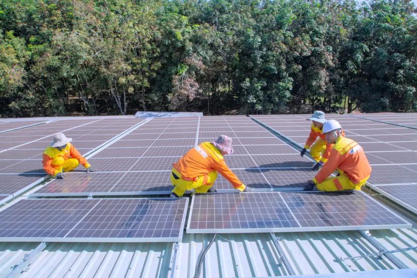 Mercado de energia com tendências e inovações no setor elétrico Quatro técnicos com uniformes de proteção amarelos e laranjas realizam a instalação e o ajuste de placas solares fotovoltaicas no telhado de uma estrutura, com uma linha de árvores verdes ao fundo