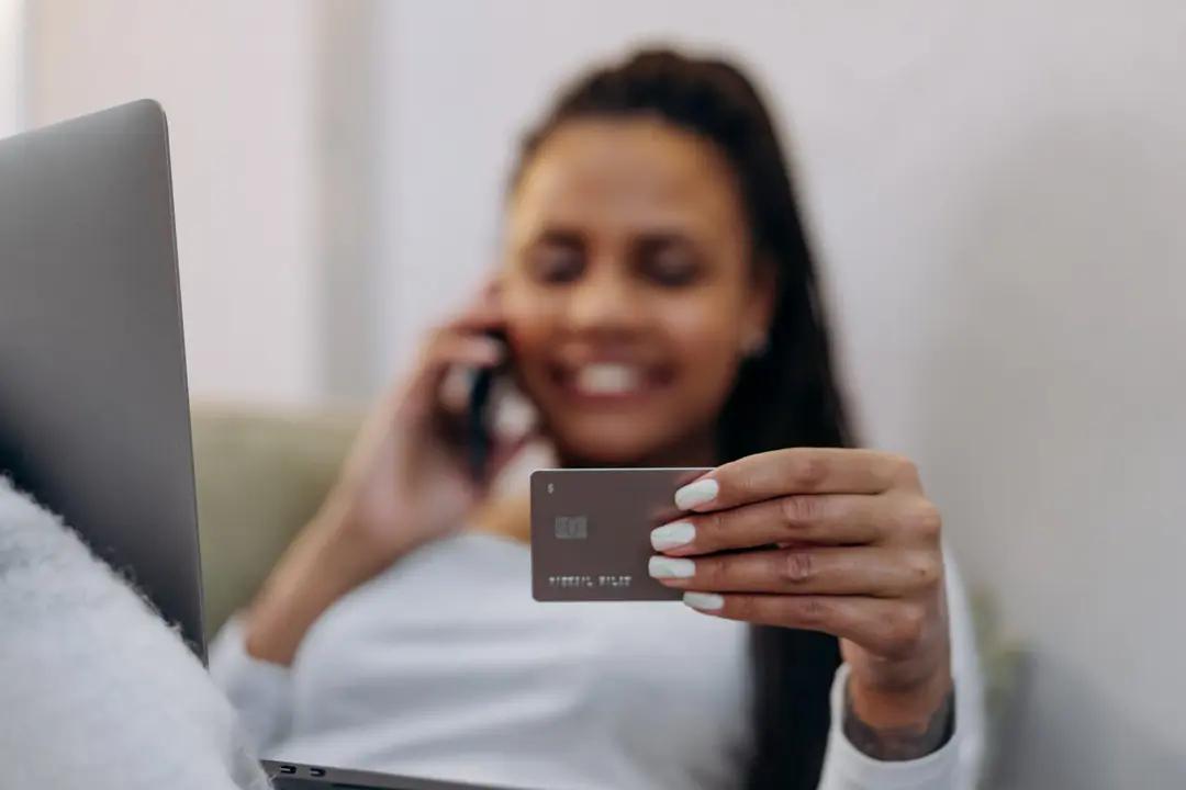 Open Finance – a evolução estratégica do Open Banking no mercado financeiro Foco na mão de uma mulher segurando um cartão de crédito cinza em primeiro plano, enquanto ela aparece desfocada ao fundo sorrindo e falando ao celular com um laptop aberto à esquerda.